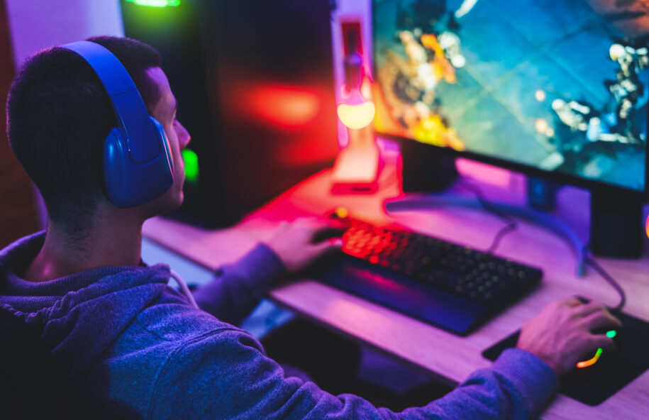 Young gamer playing online video games while streaming on social media in a dark room illuminated by RGB lights.